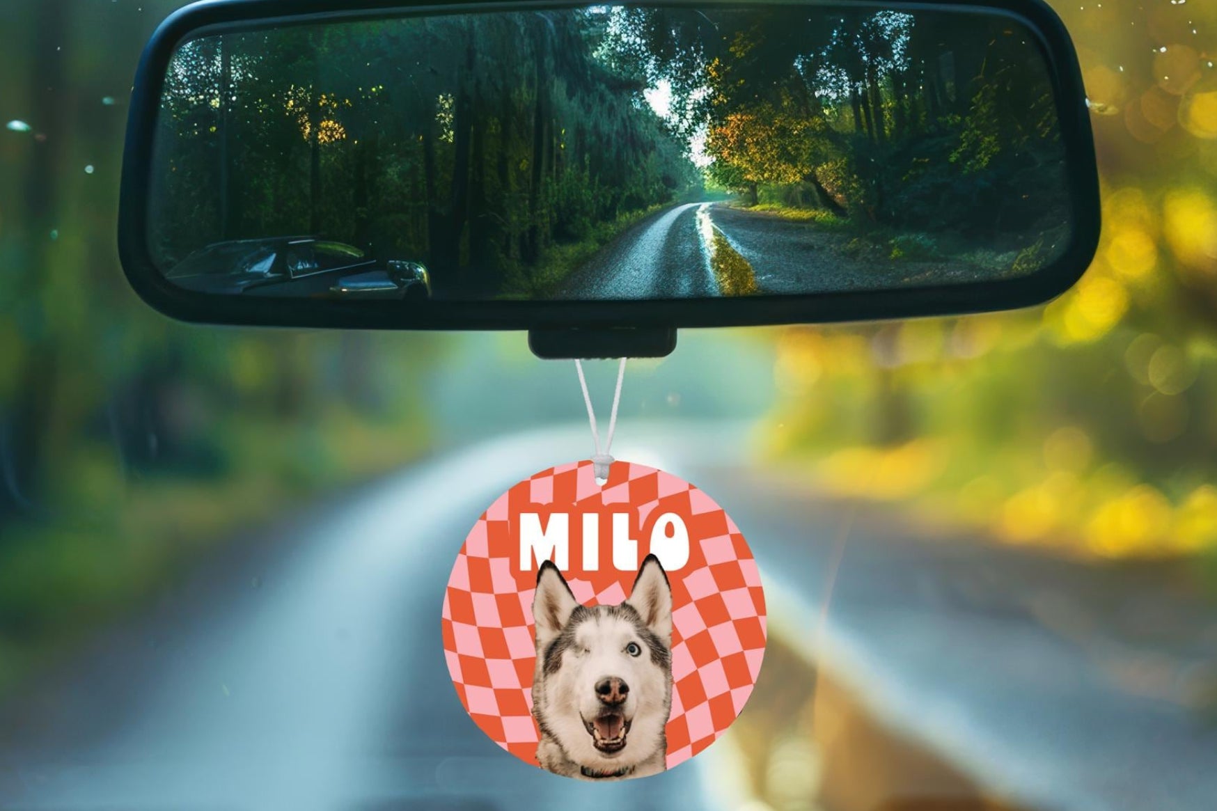 Car interior with a rearview mirror and a dog-themed air freshener hanging from it, with a blurred road and trees in the background.